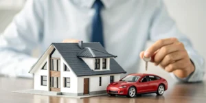 A person holds a key over a model house and car, deciding if it is better to combine home and auto insurance.